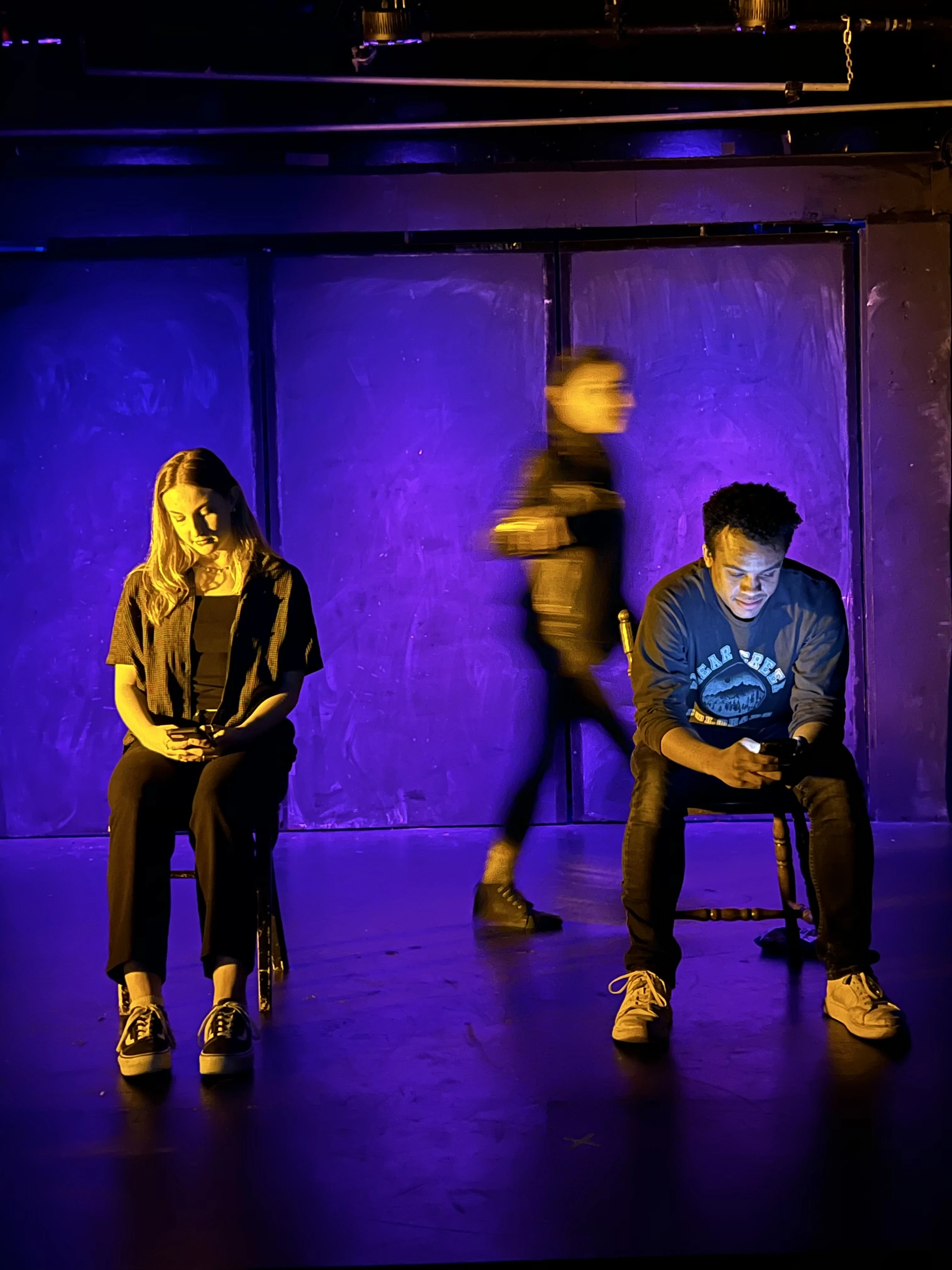 Two characters text on stage next to each other. A third walks past in dark lighting.